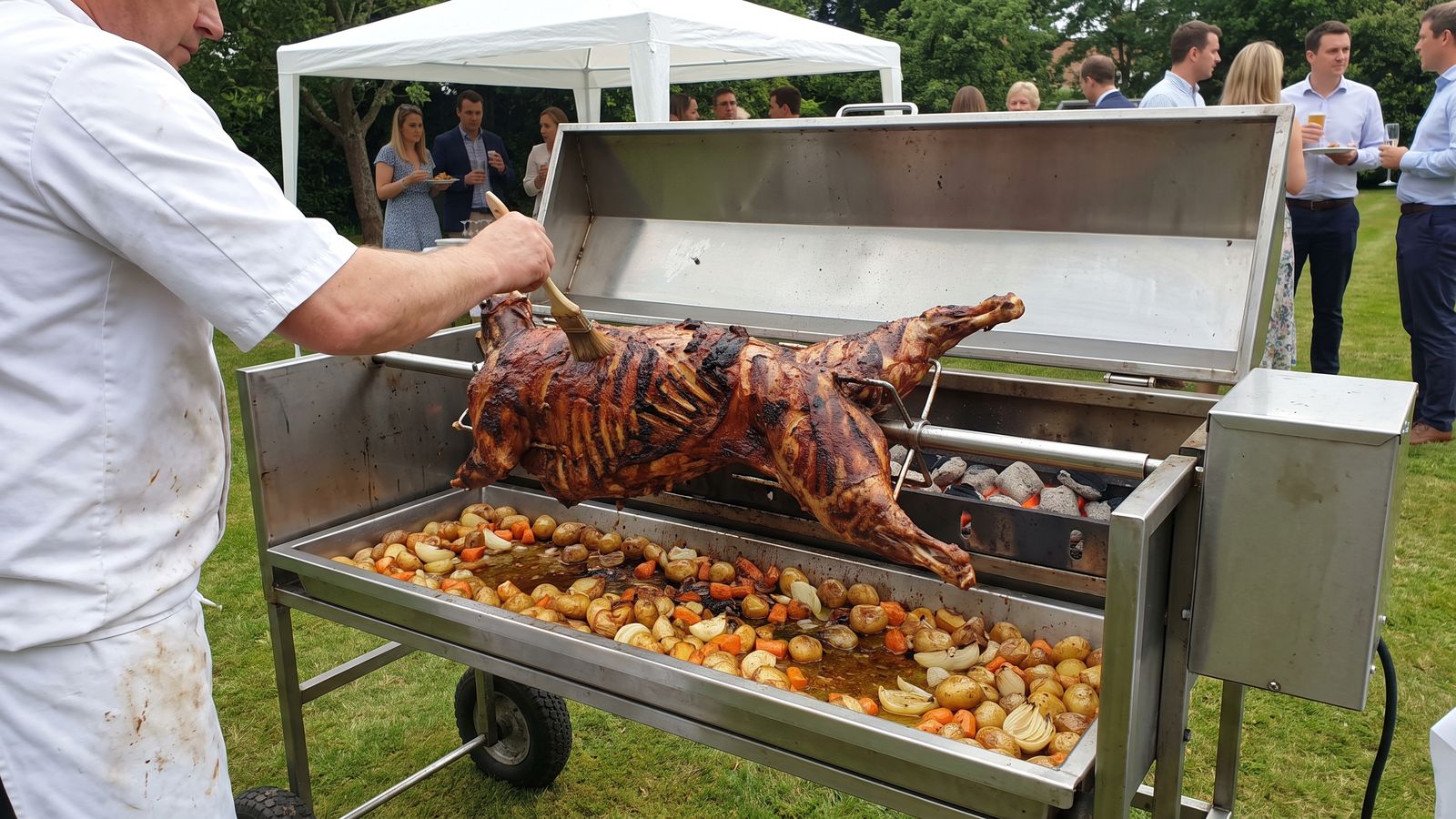 Spit braai catering in the Cape Winelands: a chef basting a whole lamb on a stainless spit machine at a garden function